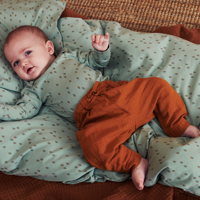 Baby dreng med mørkt hår liggende på dyne. Babyen er iført Billie baby body i støvet grøn med multifarvet bladprint og et par brune bukser.  