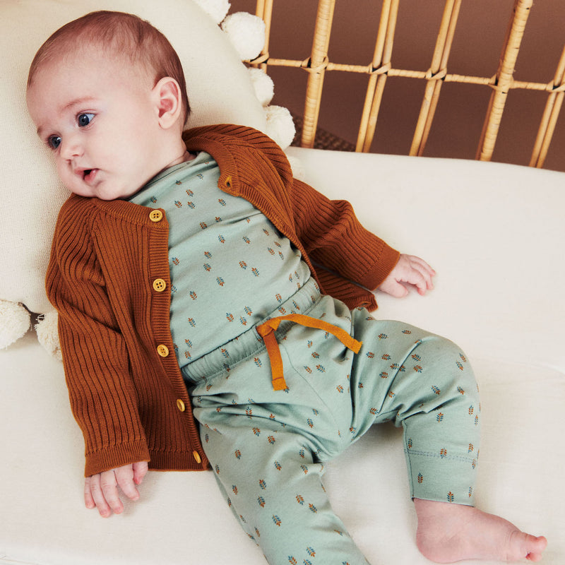 Baby med mørkt hår liggende i bambussofa med hvide hynder. Babyen er iført Popirol body og bukser i støvet grøn med multifarvet bladprint og brun strik cardigan.
