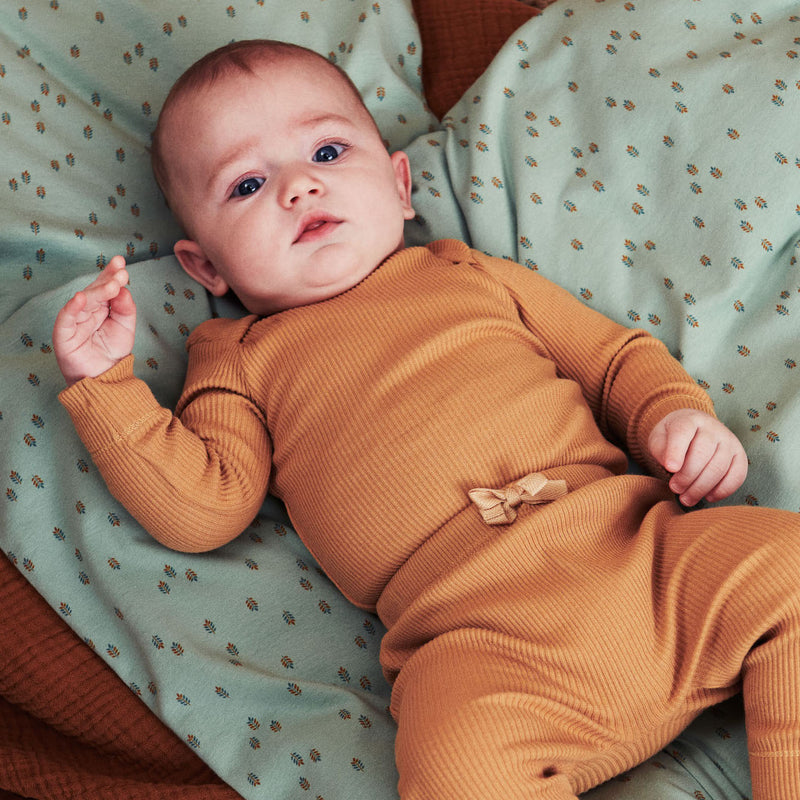 Baby dreng med mørkt hår liggende på støvet grøn dyne med multifarvet bladprint. Babyen er iført lysebrun Popirol body og bukser i rib. 