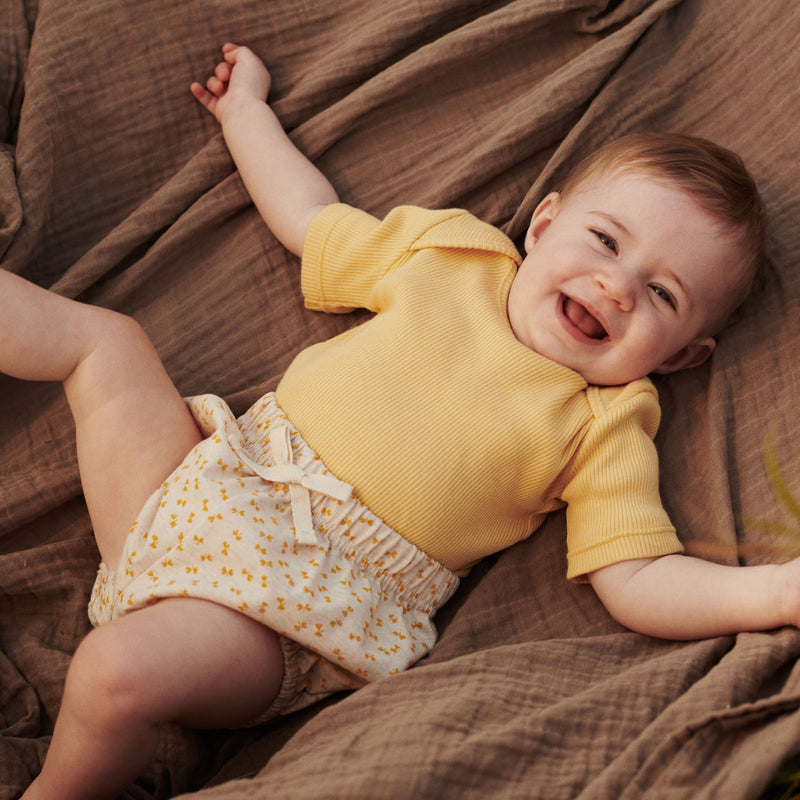 Babypige i printede bloomers og body i gule nuancer fra Popirol. Babypigen ligger på brunt tæppe med armene ud ti siderne og smiler. 