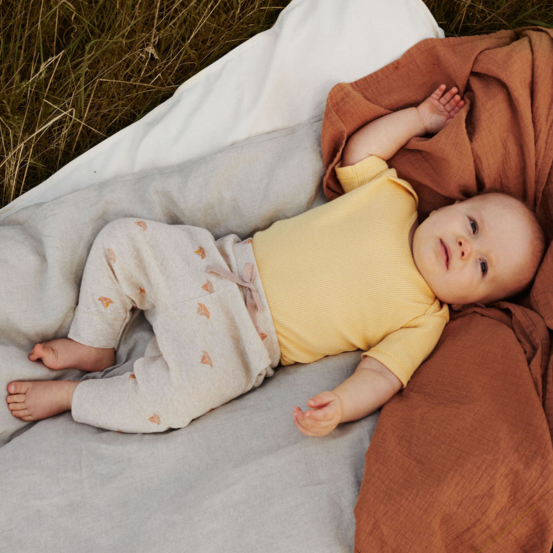 Babydreng i gul body og printede bukser fra Popirol. Babydrengen ligger på hørdyne i græsset.