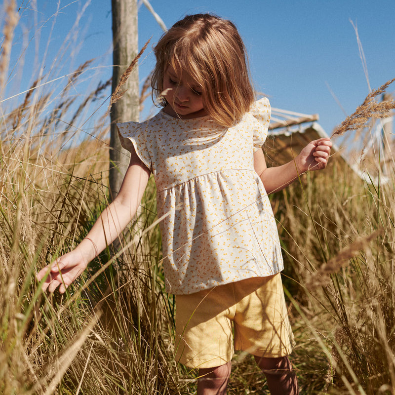 Pige i printet bluse og shorts i gule nuancer fra Popirol. Pigen står midt i en græsmark i solskin og med blå himmel.
