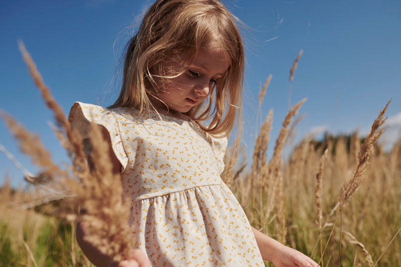 Pige i printet kjole fra Popirol. Pigen står midt i en græsmark i solskin og med vind i håret.