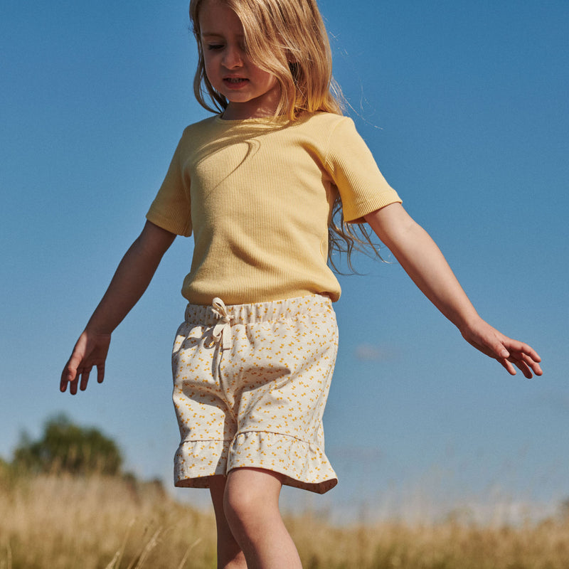 Pige i printede shorts og gul t-shirt fra Popirol. Pigen står midt i en græsmark i solskin og blå himmel.