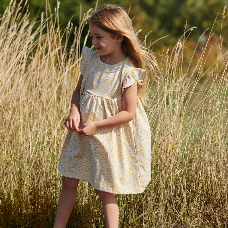 Pige i printet kjole fra Popirol. Pigen står midt i en græsmark i solskin og med vind i håret.